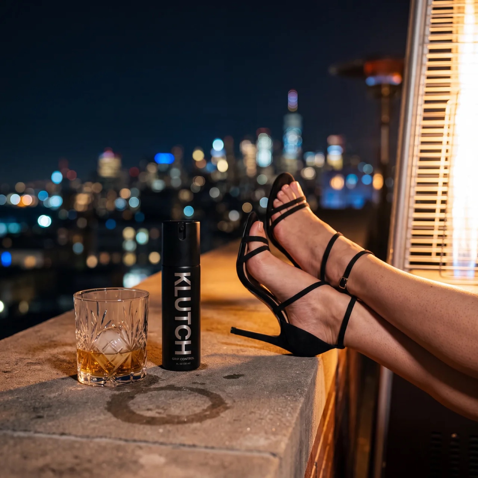 Feet in strappy heels on a rooftop ledge beside the Klutch bottle and a whiskey glass